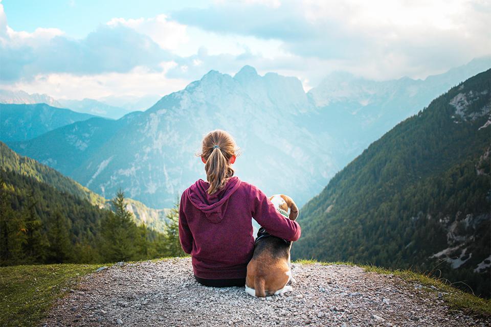 pohled ze zadu, dívka se dívá na hory a drží levou rukou svého psa kolem boku